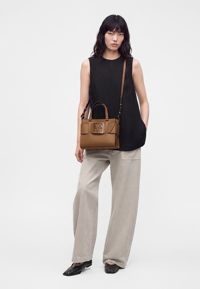 Mujer con top negro sin mangas y pantalones anchos beige, llevando un bolso de cuero marrón con una gran hebilla frontal y calzando zapatos negros.