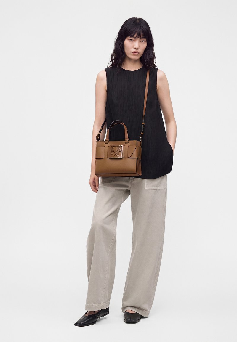Femme portant un haut noir sans manches et un pantalon beige large, portant un sac à main en cuir marron avec une grande boucle à l'avant et des chaussures noires.