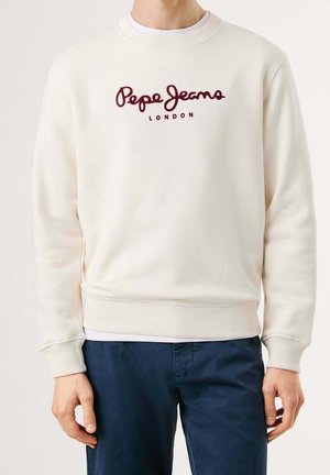 Mężczyzna w kremowej bluzie Pepe Jeans London i ciemnoniebieskich spodniach, stoi z rozluźnionymi ramionami po bokach.