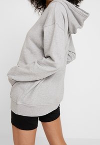 Grå bomullshoodie med en framficka, dragsko på huvan och ribbade ärmslut, bärs över svarta shorts. Mjuk textur, avslappnad design.