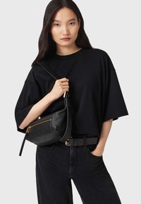Zwarte leren tas met een strak design, gouden ritssluiting en verstelbare schouderriem, gedragen met een oversized zwart t-shirt en donkere spijkerbroek.