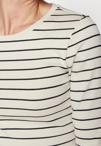 Creamfärgad topp med svarta horisontella ränder; mjukt tyg; rund halsringning; figurnära design; halsringning och ränder accentuerade med metallictråd.