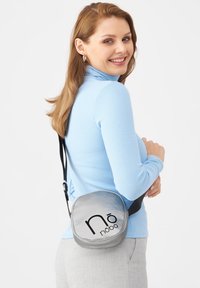 Borsa crossbody rotonda argentata con finitura lucida; presenta un logo prominente in nero, una tracolla regolabile e un design compatto.