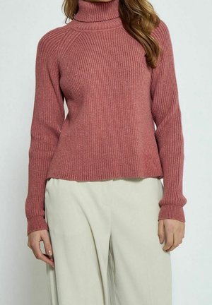 Woman wearing a rose pink ribbed turtleneck sweater paired with loose, light beige pants, standing against a plain background.