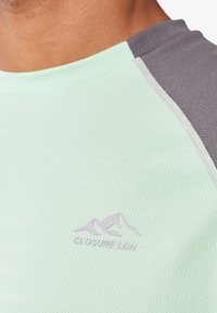 Camiseta atlética verde menta con acentos grises, que presenta un diseño de rayas texturizadas y el logo "CLOSURE LDN" en morado claro en el pecho.