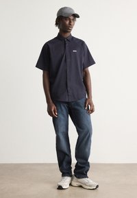 Camisa de manga curta azul-marinho com colarinho abotoado, logótipo "BOSS" bordado, combinada com jeans azuis e sapatilhas de cor clara.