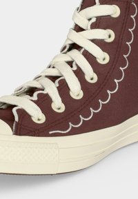 Καφέ ψηλά πάνινα παπούτσια με λευκά κορδόνια, λευκή ραφή με κυματιστό σχέδιο και κρεμ λαστιχένια σόλα και μύτη.