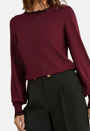 Femme portant un haut à manches longues bordeaux avec bordure noire et un pantalon noir taille haute avec boutons dorés.