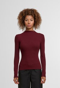 Bordó hosszú ujjú felső testhez simuló fazonnal és magas nyakkal. Simább textúra, egyszerű sziluett, látható minták vagy díszítések nélkül.