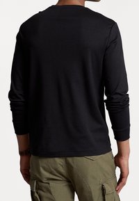 Man met een zwart shirt met lange mouwen en olijfgroene cargobroek, staand met zijn rug naar de kijker tegen een effen achtergrond.