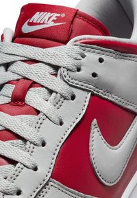 Röd och grå sneaker med läderöverdel, som har en vit Nike-logotyp, grå snörning och röda accenter på tungan och sidorna.