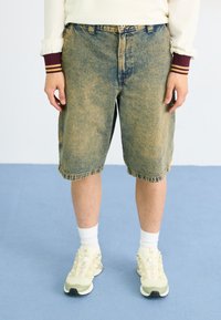 Shorts de denim con un lavado claro y textura desgastada, que presentan un corte clásico, lazos para cinturón y un dobladillo recto. Llevados con zapatos claros y calcetines blancos.