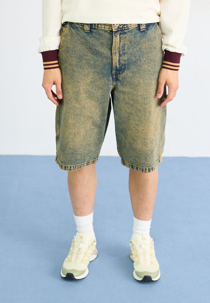 Shorts de denim con un lavado claro y textura desgastada, que presentan un corte clásico, lazos para cinturón y un dobladillo recto. Llevados con zapatos claros y calcetines blancos.