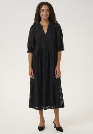 Schwarzes Spitzenkleid mit V-Ausschnitt und Dreiviertelärmeln. Hat florale Muster und eine fließende Länge bis zur Mitte der Waden. Getragen mit schwarzen Absätzen.