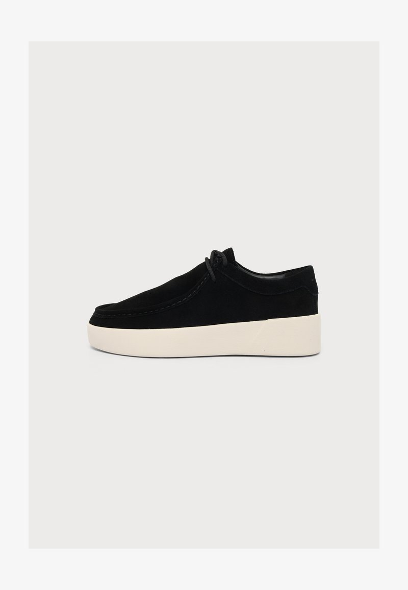 Chaussure en daim noir à lacets avec une semelle blanche épaisse, hauteur basse à la cheville et couture minimale sur un fond clair.