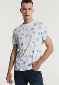 Weißes Baumwoll-T-Shirt mit einem blauen Korallen- und Seegras-Muster, rundem Ausschnitt, kurzen Ärmeln und entspannter Passform. Kombiniert mit dunklen Jeans.
