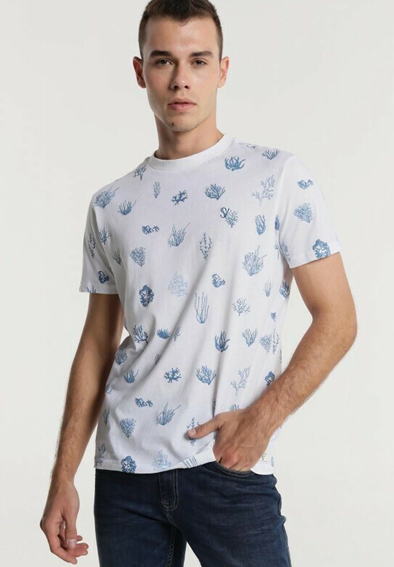 Weißes Baumwoll-T-Shirt mit einem blauen Korallen- und Seegras-Muster, rundem Ausschnitt, kurzen Ärmeln und entspannter Passform. Kombiniert mit dunklen Jeans.