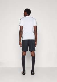 Tenue de sport comprenant un maillot blanc à manches courtes avec des accents noirs, un short noir et des chaussettes noires montant jusqu'aux genoux, conçue pour la performance athlétique.