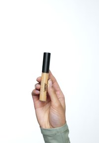 Hellbeige Concealer in einem klaren Tube mit schwarzem Deckel, in einer Hand gehalten. Mit Markenlogo in schwarzem Text an der Seite.