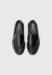 Mocassins noirs en simili cuir végétalien avec un design arrondi, dotés d'une sangle décorative et de détails de couture le long des bords. Surface supérieure texturée.