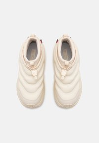 Sneakers vaaleasta beige väristä, pehmustetut ja ribbitetyllä, aaltoilevalla pinnalla. Elastinen kaulus ja vetolenkki helpottavat päälle laittoa. Kevyt kumipohja, minimaalinen muotoilu.