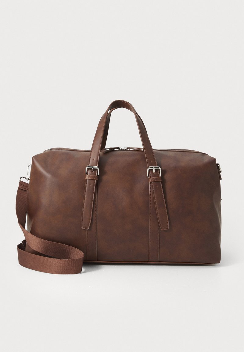Bolsa de viaje de cuero marrón con asas superiores, correa desmontable, cierre de cremallera y textura elegante. Diseño minimalista sin logotipos visibles.