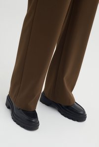 Schwarze klobige Slipper mit einer strukturierten Oberfläche, kombiniert mit braunen weit geschnittenen Hosen aus einem glatten Stoff, sichtbare Nahtdetails.
