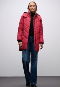 Abrigo acolchado rojo con capucha, con cierre de cremallera en la parte delantera y dos bolsillos laterales. Combinado con jeans de mezclilla azul de pierna ancha.