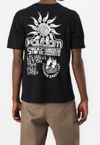 Camiseta negra de manga corta con diseños gráficos en blanco, incluyendo un motivo de sol y texto con nombres de ciudades y detalles del evento. Material de algodón.