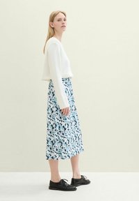 Frau in weißem Pullover, blauem Blumen-Midi-Rock und schwarzen Schnürschuhen, die vor einem hellen neutralen Hintergrund steht.