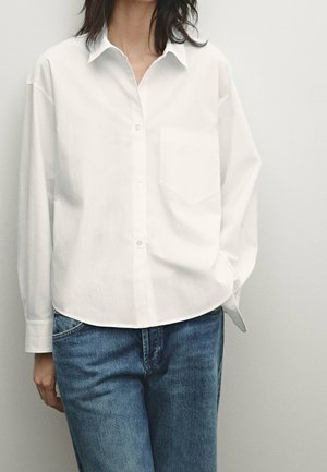 Femme portant une chemise blanche ample boutonnée avec poche poitrine et un jean bleu, debout devant un fond clair uni.