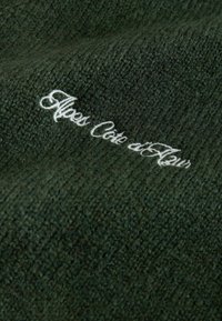 Donkergroene gebreide stof met een zachte textuur en witte geborduurde tekst waarop staat: "Alpes Côte d'Aleur."