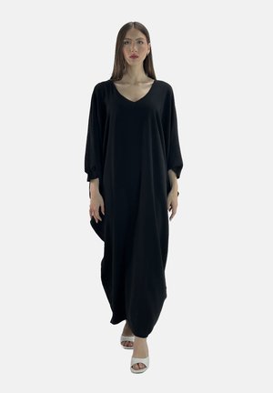 Abito nero oversized con scollatura a V profonda, maniche ampie e silhouette fluida. Il modello indossa sandali con tacco bianchi. Design semplice e monocolore.