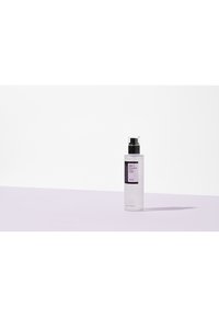 Bouteille en plastique transparente avec un capuchon-pompe noir ; l'étiquette présente un texte noir sur fond blanc ; le contenu est visible ; positionnée sur une surface mauve clair.
