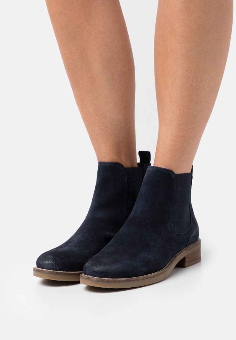 Bottines en daim bleu marine avec un petit talon marron et des panneaux latéraux élastiques, portées sur jambes nues sur fond blanc.