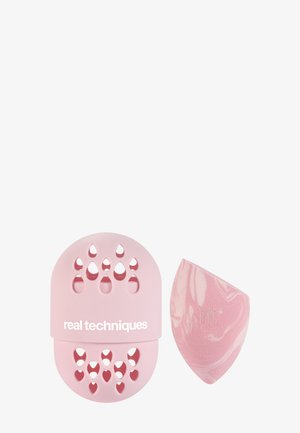 Pink makeupsvamp og matchende silikoneholder med dråbeformede udskæringer og "real techniques" branding på en hvid baggrund.