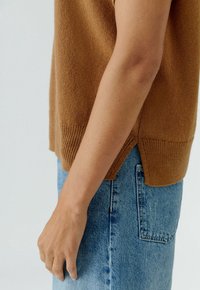 Bruine gebreide trui met geribbelde onderrand, gedragen over lichtblauwe denim. De trui heeft zijsplitten, waardoor een ontspannen pasvorm en zachte textuur zichtbaar zijn.