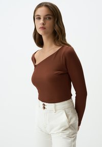 Braunes geripptes Langarmoberteil mit diagonaler Ausschnittlinie und goldfarbener Knopfdekoration, kombiniert mit hochtaillierten weißen Hosen mit doppelten Knöpfen.