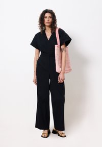Zwarte jumpsuit met korte mouwen en wijde benen; het model houdt een roze gebreide tas vast en draagt zwarte sandaaltjes met bandjes.