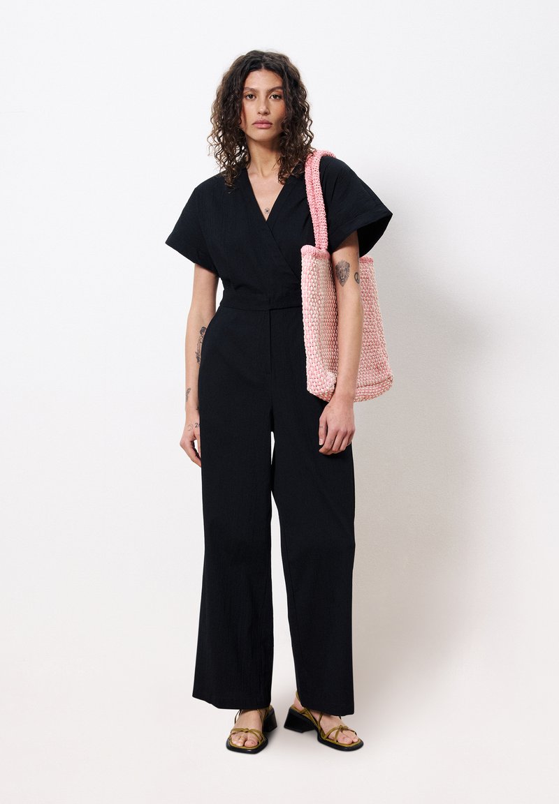 Zwarte jumpsuit met korte mouwen en wijde benen; het model houdt een roze gebreide tas vast en draagt zwarte sandaaltjes met bandjes.