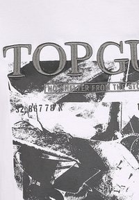 Vit T-shirt med en grafisk design i svart och grått. Texten lyder "TOP GUN" ovanför "INGEN SKYDD FRÅN STORMEN" med abstrakta mönster.