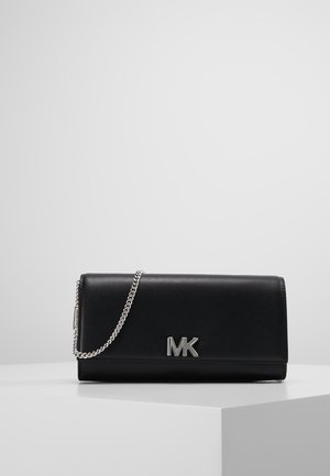 Zwarte leren Michael Kors clutch met zilveren kettingriem en zilveren MK-logo aan de voorkant, uitgestald op een wit voetstuk tegen een grijze achtergrond.