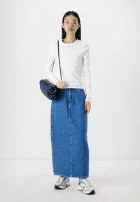 Camisetta bianca a maniche lunghe, gonna maxi in denim blu e sneakers argentati. Una borsa blu navy appoggia sulla gonna. Design minimalista, tonalità neutre.