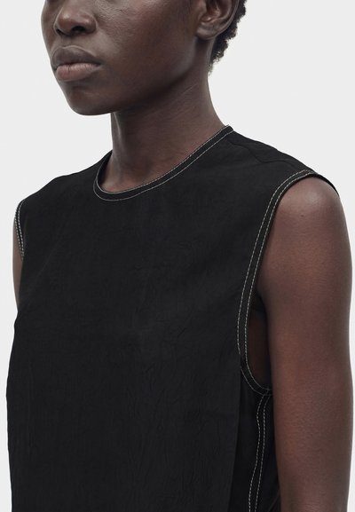 Κοντινό πλάνο ενός ατόμου που φοράει αμάνικη μαύρη μπλούζα με λευκό ραφές αντίθεσης κατά μήκος των άκρων, μπροστά σε απλό φόντο.