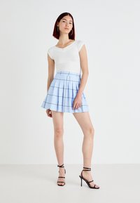 Top bianco a maniche corte abbinato a una gonna a pieghe blu a quadri. La modella indossa sandali neri con cinturini e tacco alto. Motivi semplici, linee pulite.