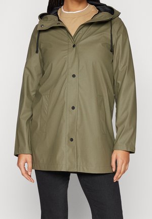 Persona con un chubasquero verde oliva con capucha y botones negros, bolsillos delanteros, suéter beige y pantalones negros sobre un fondo liso.