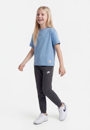 Uśmiechnięte dziecko idzie, nosi jasnoniebieską koszulkę Nike, ciemnoszare spodnie dresowe Nike oraz białe buty Nike z granatowym logo.