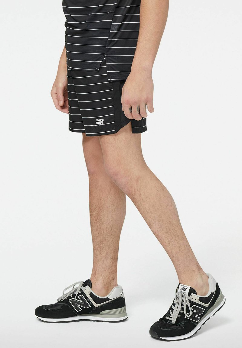Mann trägt schwarze New Balance Laufschuhe, schwarze Shorts und ein schwarzes kurzärmliges Hemd mit weißen horizontalen Streifen, steht vor einem weißen Hintergrund.