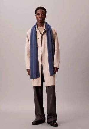 Jonge man met een beige jas, een blauwe sjaal, donkere broek en zwarte schoenen staat tegen een effen grijze achtergrond.