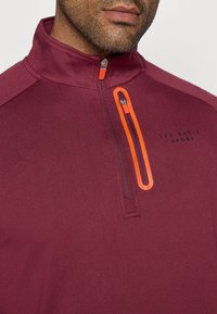 Burgundia spordipullover kõrge kraega, neljanda osa tõmblukuga ja erksast oranžist tõmbluku aktsendiga. Omab kangast tekstuuri ja logo detaile.